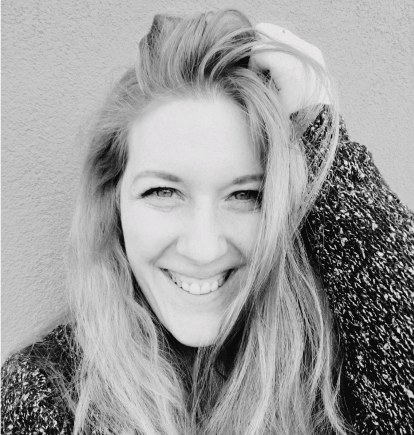 Black and white photo of a person smiling at the camera. They have long hair and are wearing a textured sweater. Their hand is placed on the top of their head, slightly tousling their hair.