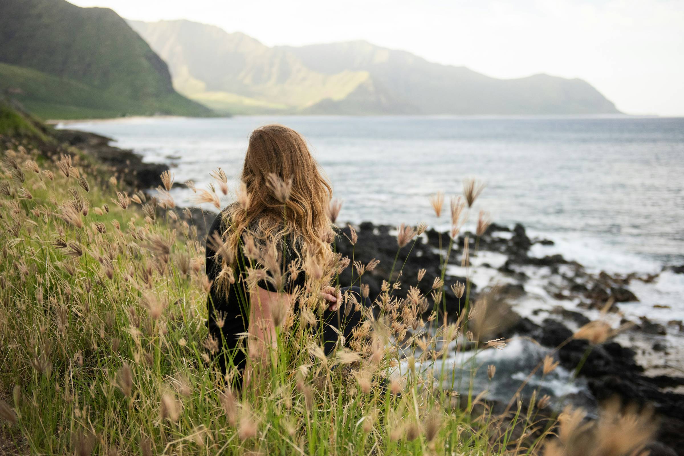 woman from the back sitting on the coast of the beach surrounded by grass and hills
