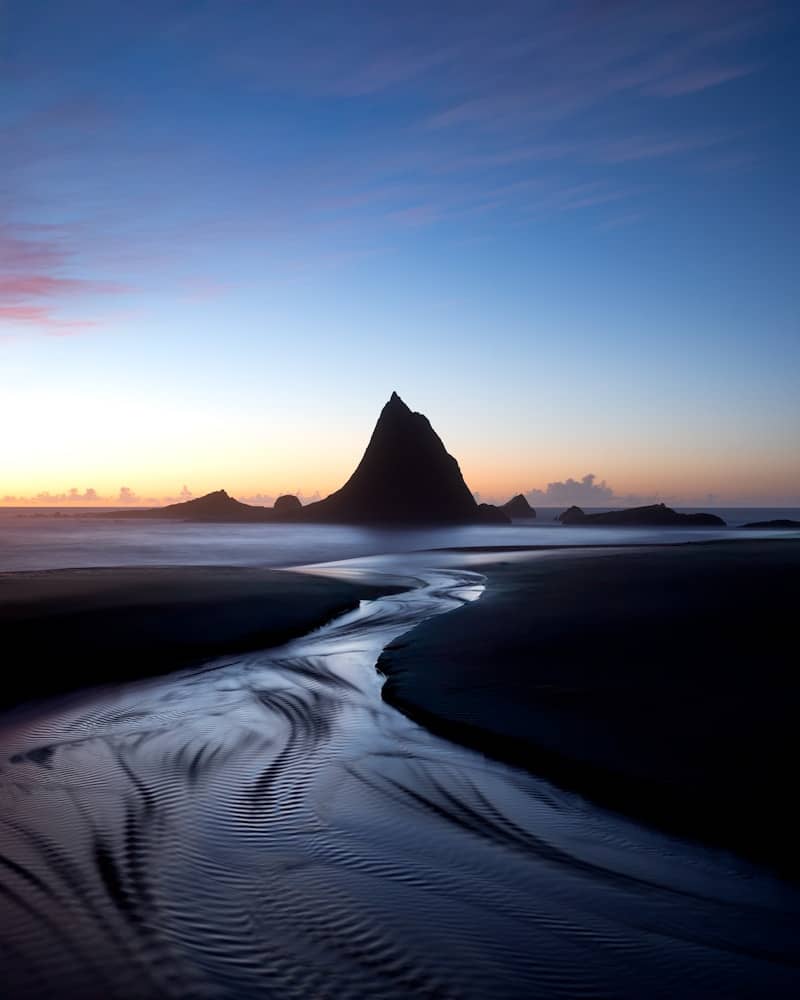 Dark beach stream flowing towards a prominent peak at dusk.