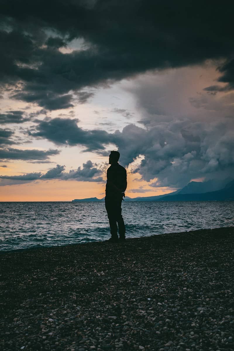 A person in silhouette on a pebble beach against a dramatic cloudy sky at low sun