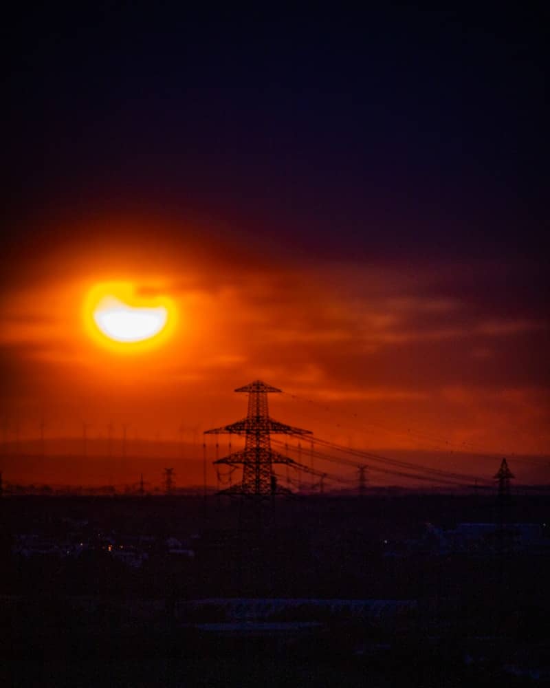 A partial solar eclipse above a silhouette of electrical transmission towers at low sun