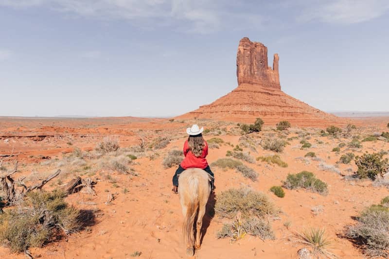Woman in cowboy hat rides horse in desert landscape.