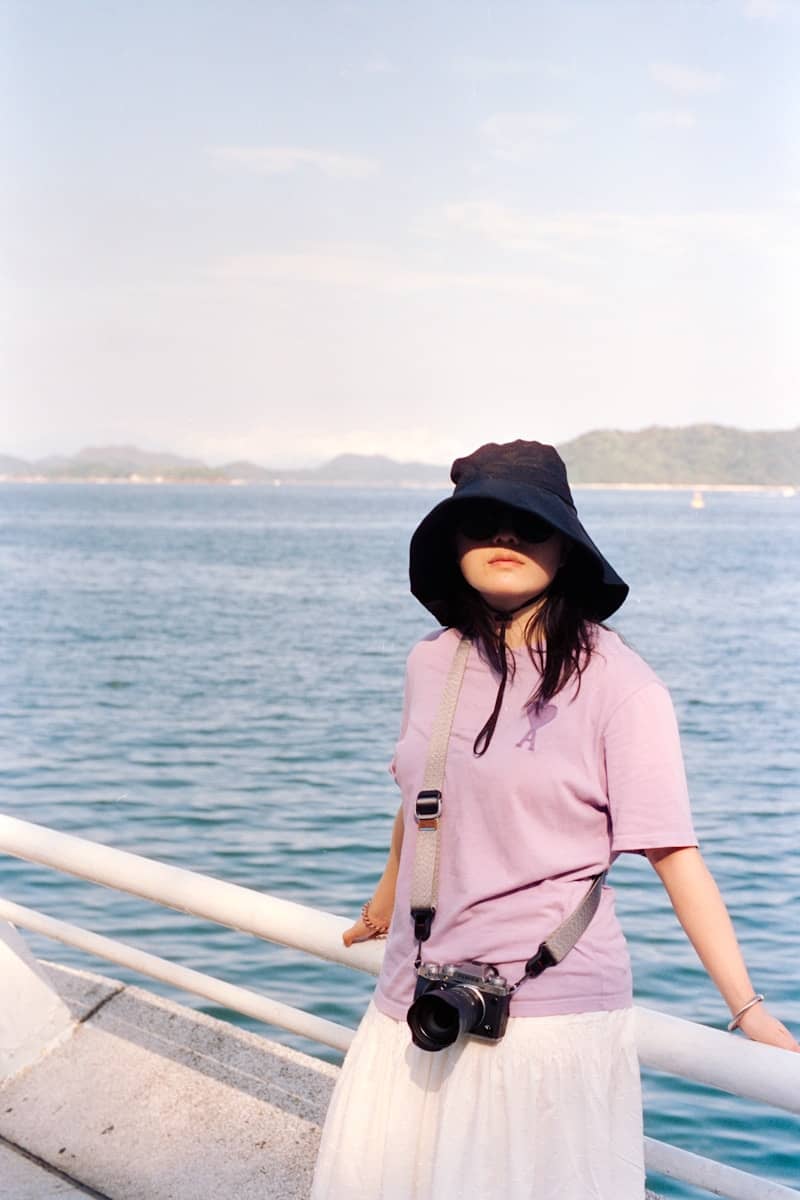 Woman with hat and camera poses by the ocean.