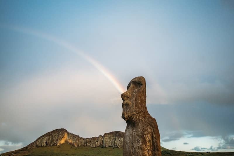 Rainbow arches from a moai statue against a cloudy sky