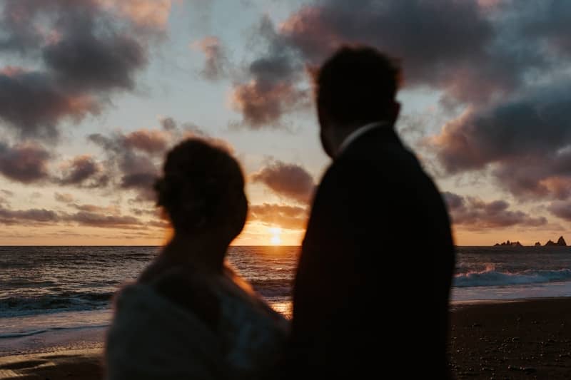 a bride and groom standing on the beach at sunset