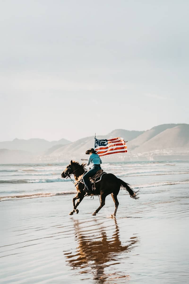 Cowgirl rides horse with american flag on beach.
