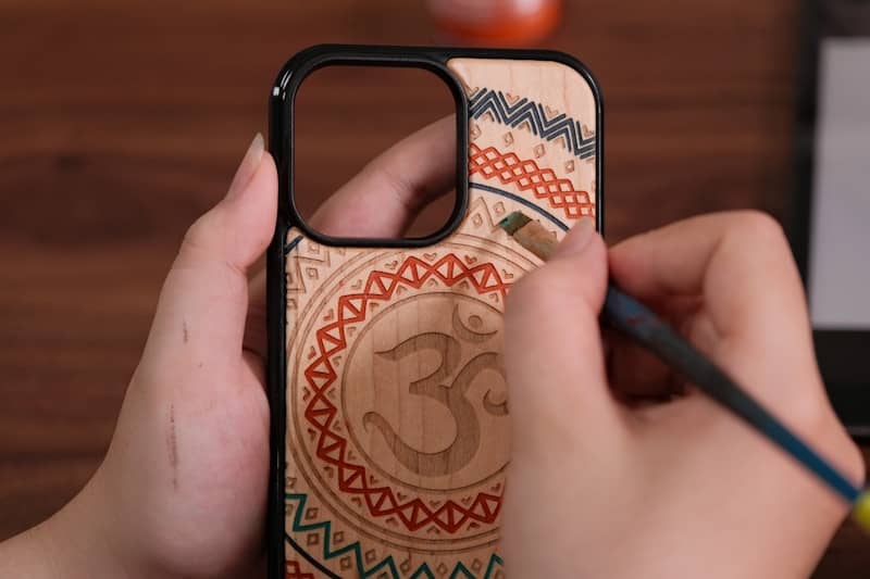 Hands decorating a wooden phone case with an om symbol.