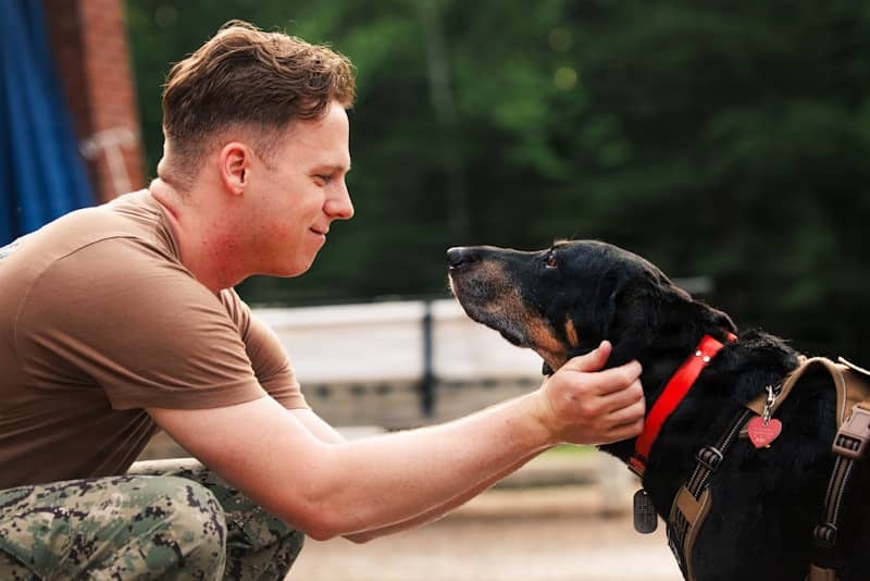 Man in military uniform petting a black dog outdoors.