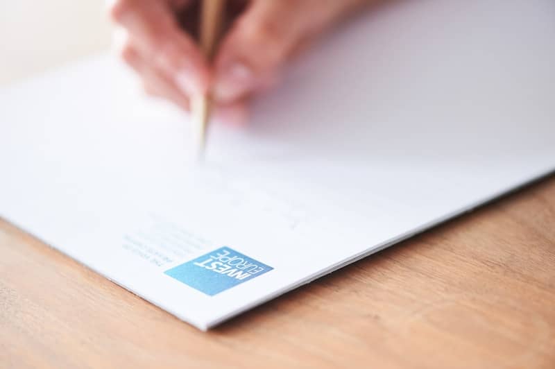 A hand writes on white paper with a blue logo.