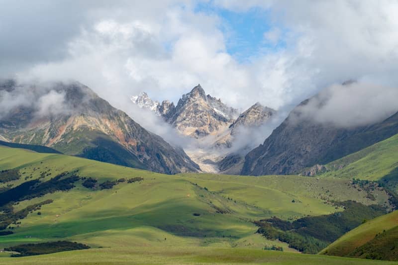 Green hills lead to majestic mountains partially covered by clouds.