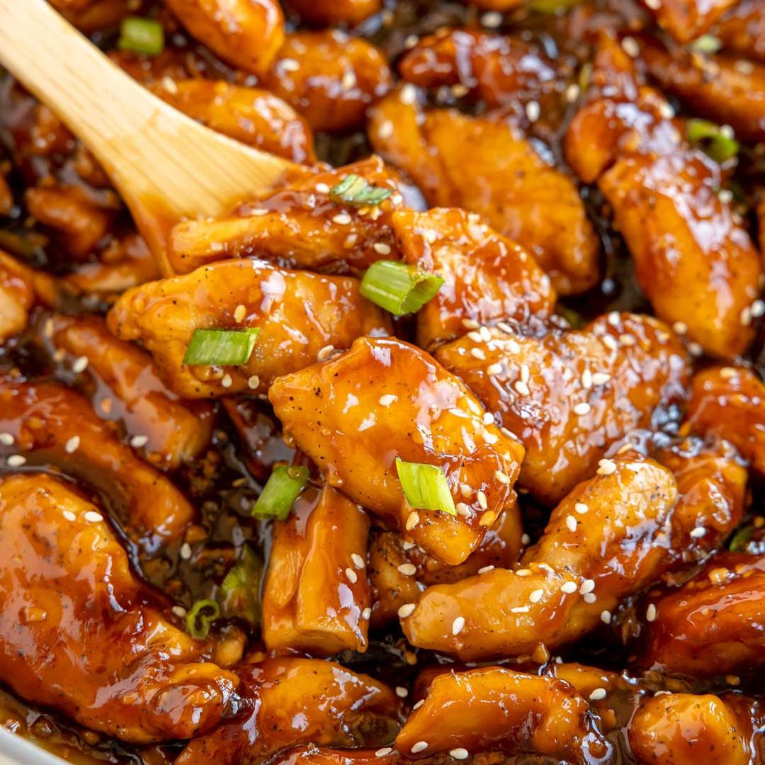 Close-up image of glazed chicken pieces coated in a rich, shiny sauce, garnished with sesame seeds and sliced green onions. A wooden spoon is partially visible, mixing the dish, showcasing the juicy and tender appearance of the chicken.