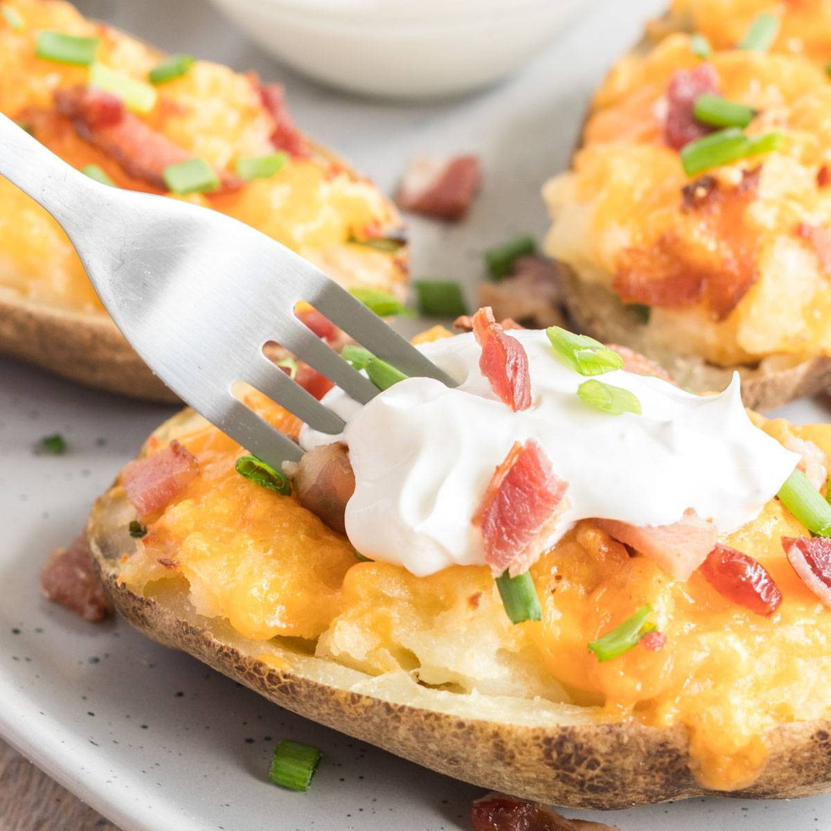 A close-up of a baked potato half topped with melted cheese, crumbled bacon, chopped green onions, and a dollop of sour cream. A fork is poised to take a bite. Fresh ingredients and vibrant colors make the dish appear appetizing. Another potato half is partially visible in the background on a speckled plate.