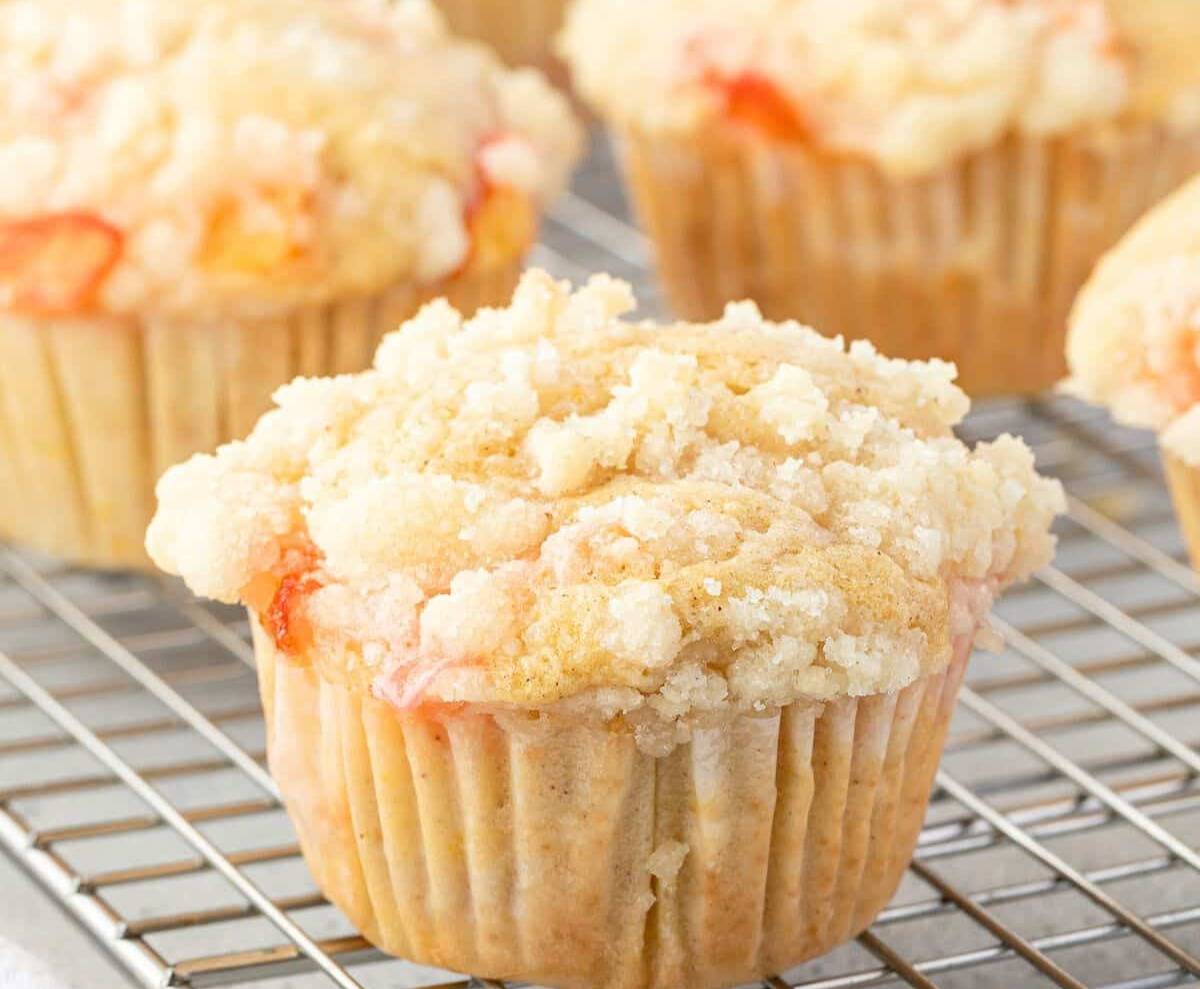 Freshly baked peach muffins cooling on a wire rack with a basket of peaches and jug of milk in the background.