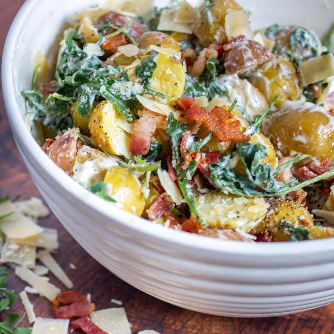 A white bowl filled with a creamy salad featuring halved potatoes, leafy greens, crispy bacon pieces, and shredded cheese. The salad is well-mixed, with visible seasoning and dressing, and a few cheese shavings are scattered on the wooden surface next to the bowl.