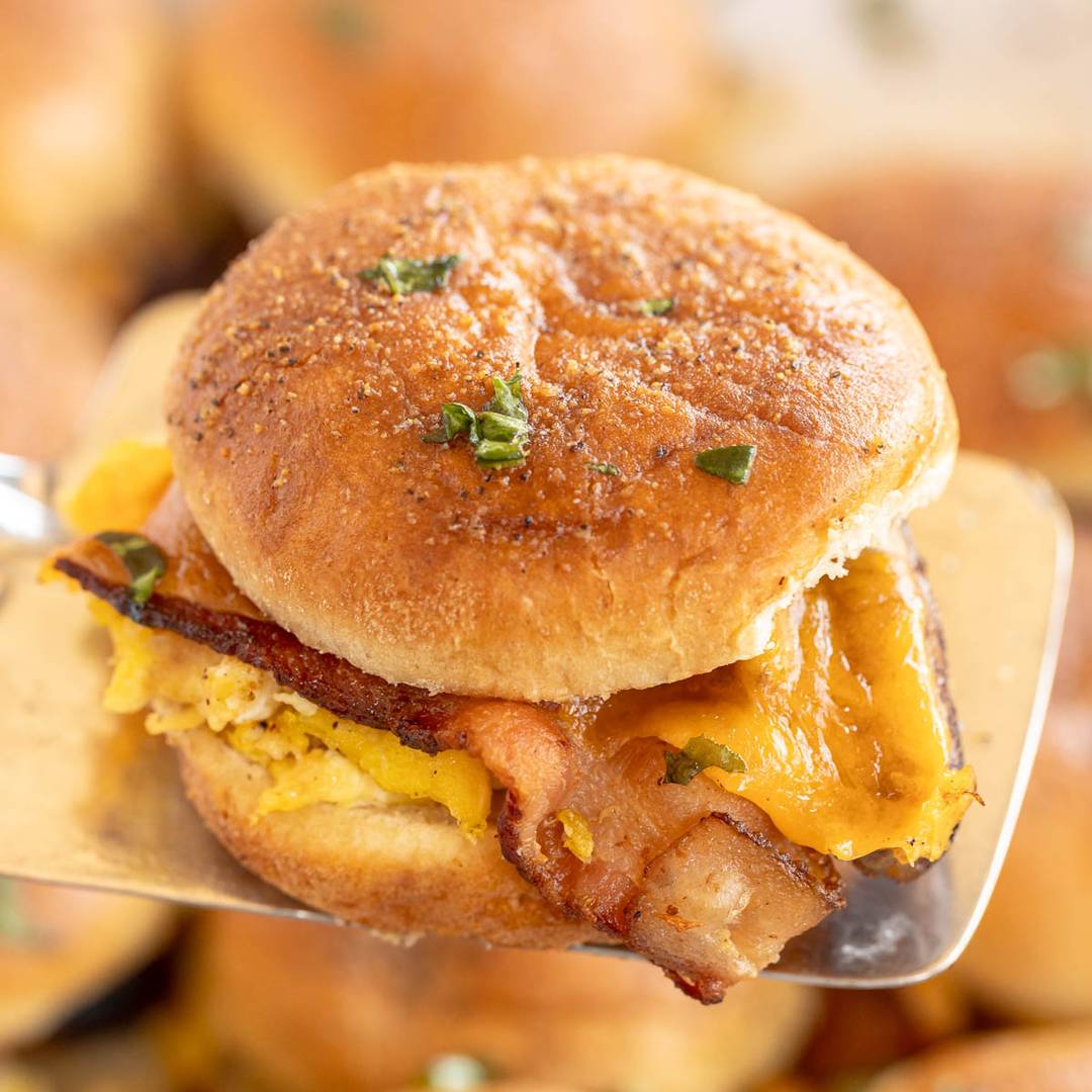 A close-up of a breakfast sandwich being served on a spatula. The sandwich consists of a soft, golden brown bun sprinkled with herbs. Inside, there are layers of crispy bacon, fluffy scrambled eggs, and melted cheese. The background features more sandwiches out of focus.