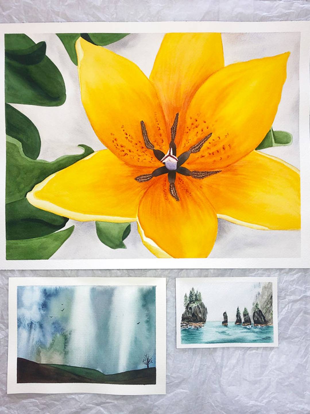 three watercolor paintings in three sizes top is an orange flower, bottom left a rainy sky, bottom right rocks in the ocean