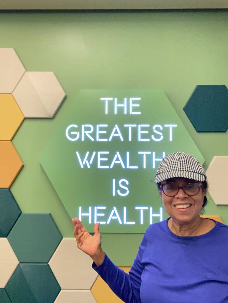 A person wearing glasses and a checkered cap is smiling and gesturing with one hand in front of a wall. The wall features hexagonal tiles in various colors and a neon sign that reads, 