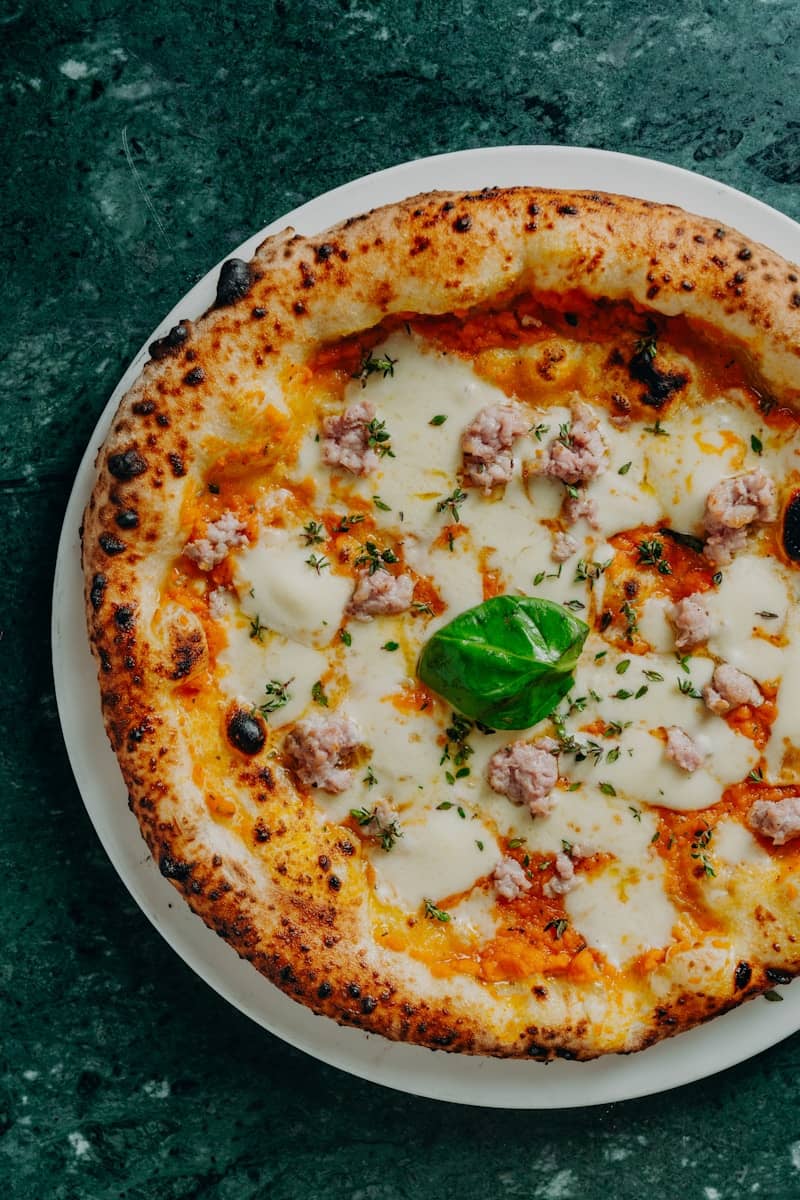 A pizza with sausage, mozzarella, and fresh basil on a dark green marble surface