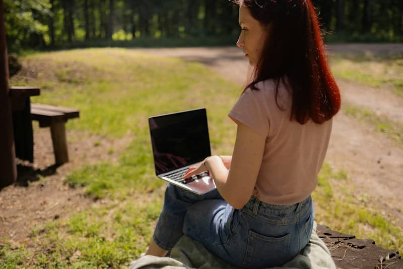 A woman with red hair typing on a laptop in a sunlit forest