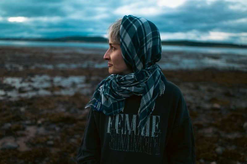 A person wearing a plaid headscarf and a dark sweatshirt on a rocky beach