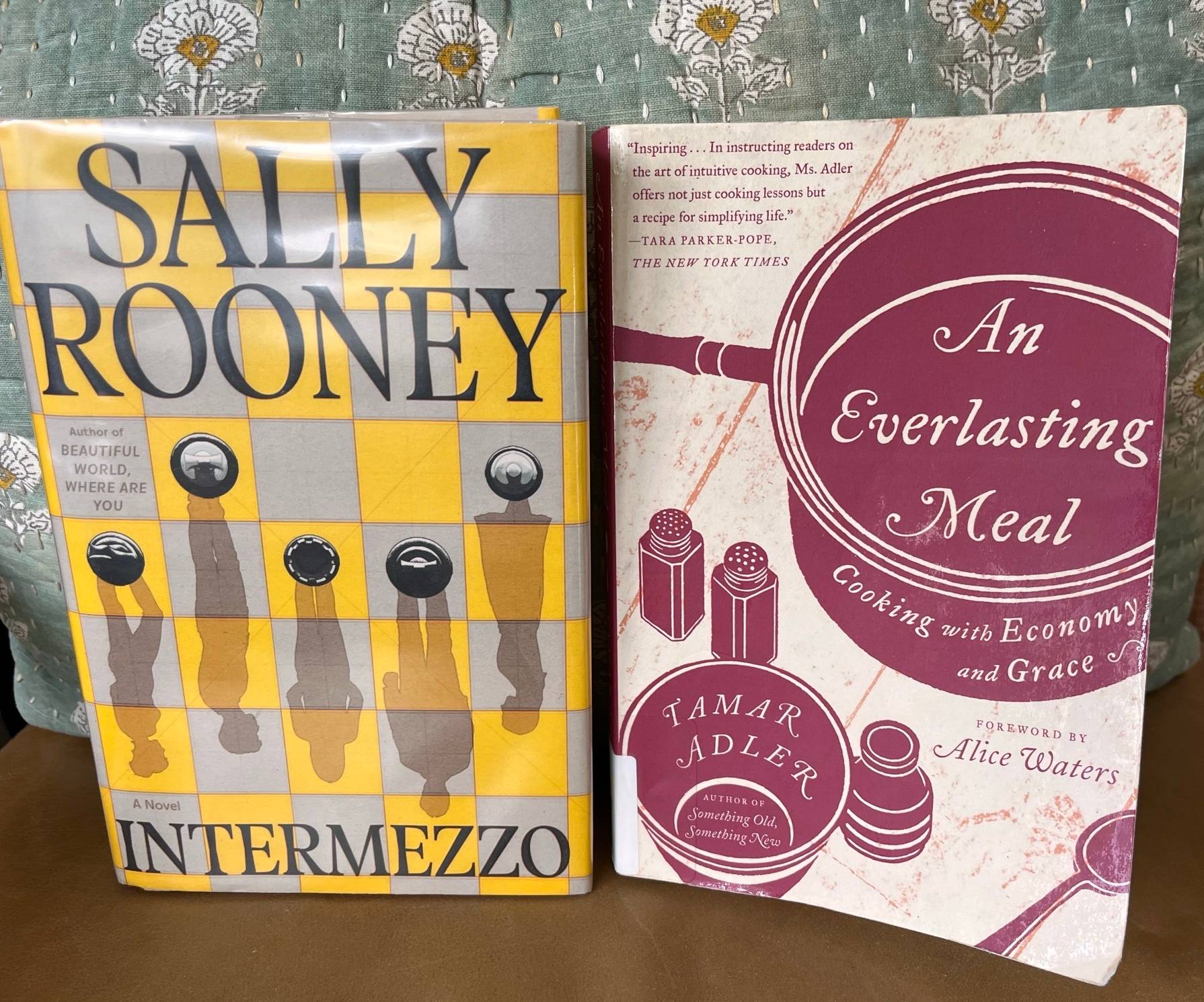 Two books, Rooney's with a grey and yello chessboard with four pieces, their shadows figures. The second book depicts a pot, salt and pepper shakers, bowl, and spoon on a table. Both books are standing up against a green pillow with white flowers on a leather chair.