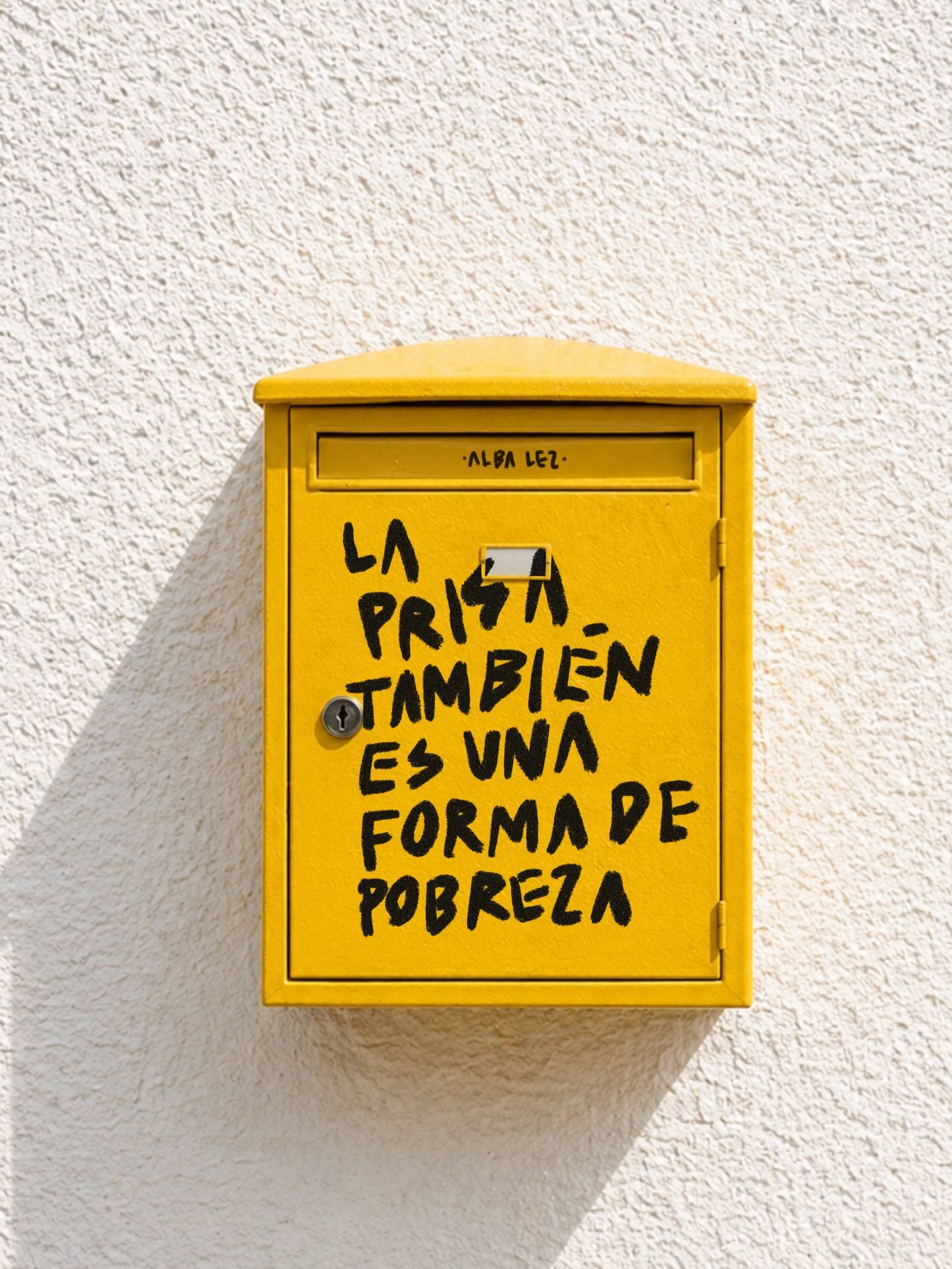 A yellow mailbox with a text in it that says "La prisa también es una forma de pobreza", or "Hustle is also a form of poverty" in English.