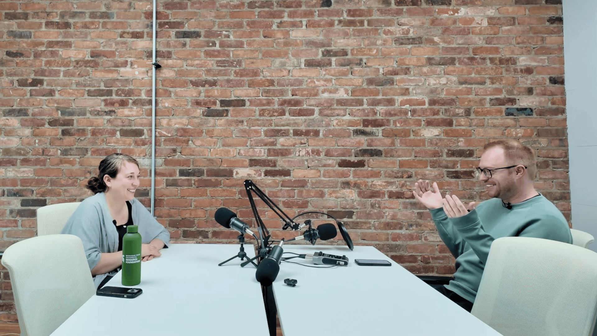 Two people sitting at a white table during a podcast recording in a brick-walled studio. On the left, a woman in a light blue cardigan holds a green water bottle, and on the right, a man in a green sweatshirt gestures while speaking. Professional micropho