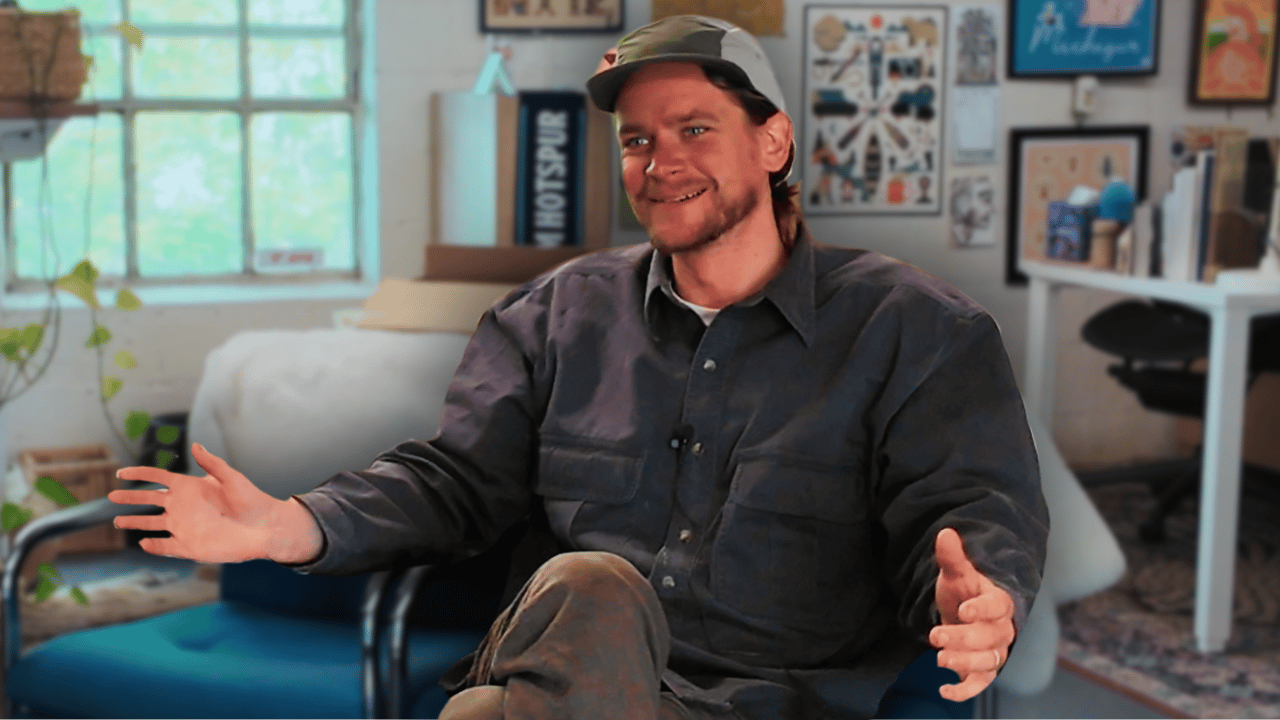Kylen Blom sitting on a couch in his bright, art-filled studio space, wearing a dark button-up shirt and baseball cap, gesturing while speaking during an interview