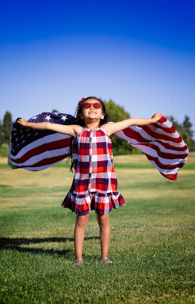 Young girl in plaid dress holding american flag like wings.