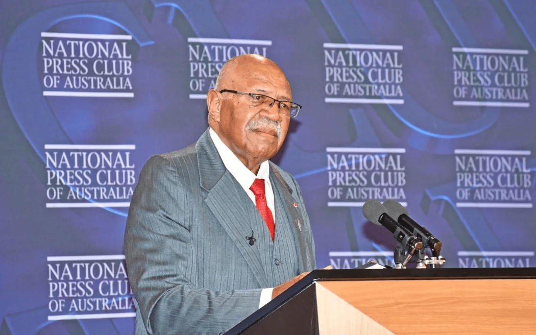 A man in a gray suit and red tie, Fiji's Prime Minister Sitiveni Rabuka, speaking at a wooden podium with multiple microphones. Behind him is a blue backdrop with repeated National Press Club of Australia logos. He is wearing glasses and has a mustache.