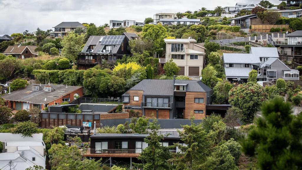 A hillside residential neighborhood with various modern houses built on a steep slope. The homes feature different architectural styles, including some with solar panels on their roofs. Lush green vegetation and trees surround the properties, creating a v