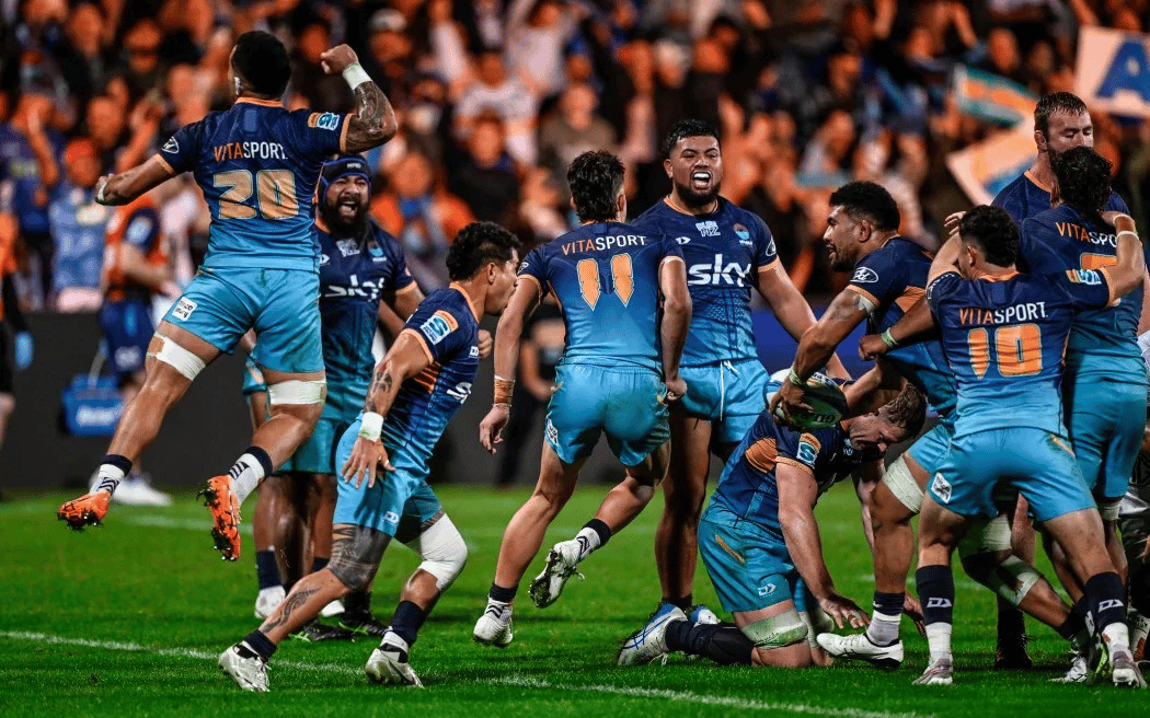 Moana Pasifika players in blue and teal jerseys celebrate enthusiastically on the rugby field, with players jumping and embracing after scoring, while crowds in orange fill the stadium stands in the background.
