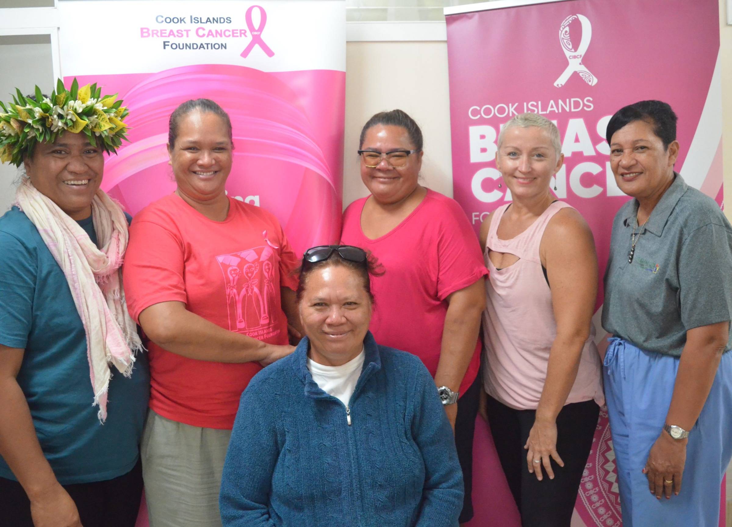 A group of six women, mostly of Pacific Islander descent, are posing for a photo indoors. Five women are standing, and one is seated in front. Behind them are two pink banners with Cook Islands Breast Cancer Foundation and a pink ribbon logo. Most of the women are smiling and wearing pink or light-colored clothing. One woman on the left wears a floral headpiece.