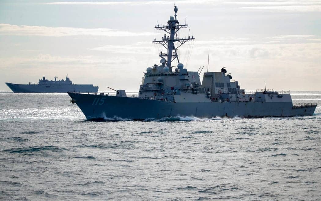 A large grey warship, possibly a destroyer with the number 115 visible on its bow, is cutting through the water, creating a wake. In the background, another larger grey ship with a flat deck is visible on the horizon under a cloudy sky.