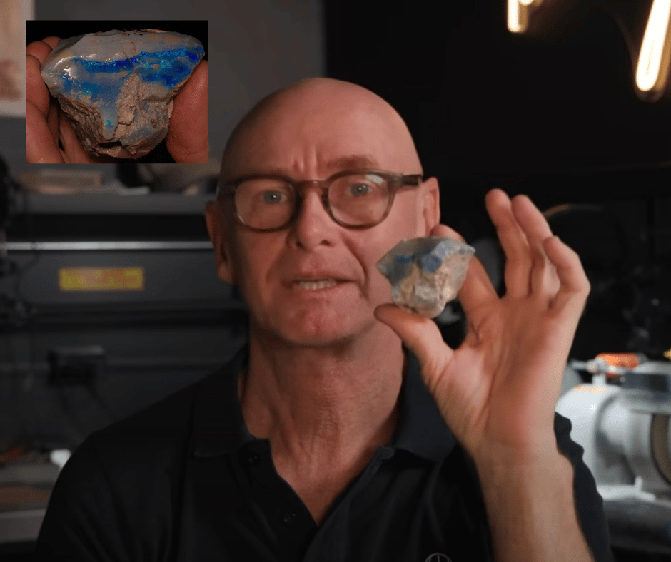 A bald man wearing glasses, Justin Thomas, holds up a raw opal stone showing brilliant blue and green colors against brown rock matrix. In the background, a computer monitor displays a close-up image of the same opal specimen, revealing the stone's vibrant play of color in greater detail.
