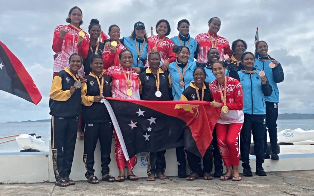 Women's medal-winning teams from Papua New Guinea, Tonga, and Fiji pose together at the 2025 Palau Games after a va'a (outrigger canoe) race. Athletes wear their national uniforms and proudly display gold, silver, and bronze medals, with the PNG team holding their national flag in front. The ocean and canoes are visible in the background under an overcast sky.