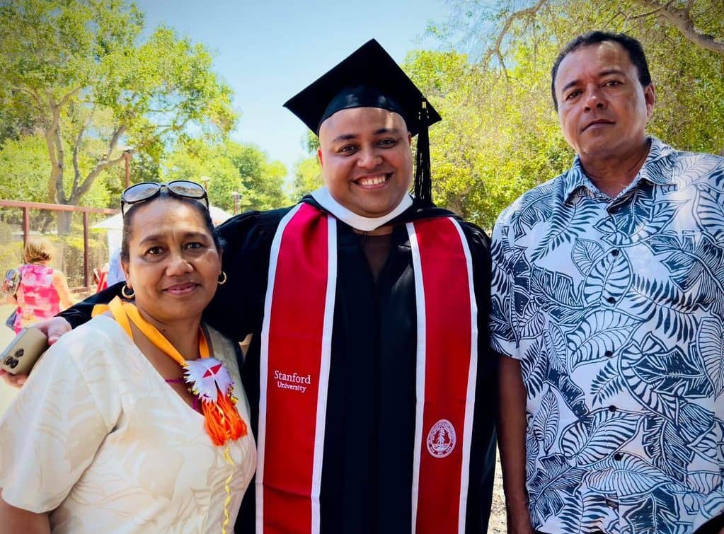 Wilfred Singh Fimone celebrates his Stanford University graduation with his parents in an outdoor ceremony setting. He stands in the center wearing traditional black graduation cap and gown with a distinctive red and white Stanford University hood, smiling broadly at the camera. His mother stands to his left wearing a white top with a colorful floral lei, while his father stands to his right wearing a blue and white tropical print shirt. The scene is set against a backdrop of lush green trees and clear blue sky, capturing a proud family moment at the Stanford campus graduation ceremony where Fimone became possibly the first Rotuman to earn a Master's degree in Linguistics from the prestigious university.