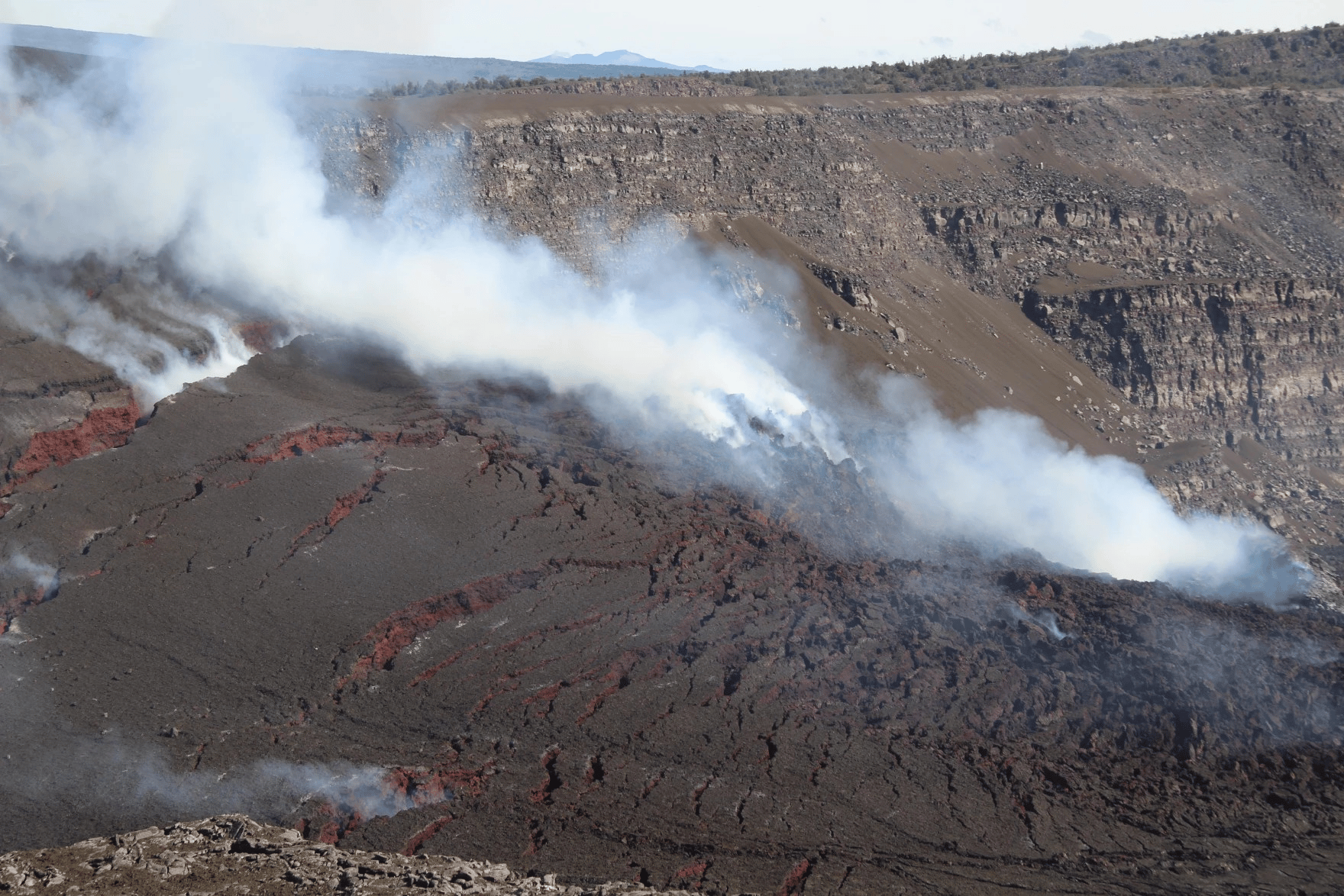 A dramatic wide shot captures a volcanic caldera with an active lava flow and plumes of smoke. The foreground shows a large expanse of dark, solidified lava, with fissures revealing bright orange and red molten lava glowing beneath the surface. Multiple streams of white and grey smoke rise from various points on the lava field, obscuring parts of the view. The background features steep, rugged caldera walls in shades of brown and grey, topped by a distant, hazy horizon of hills under a light sky.