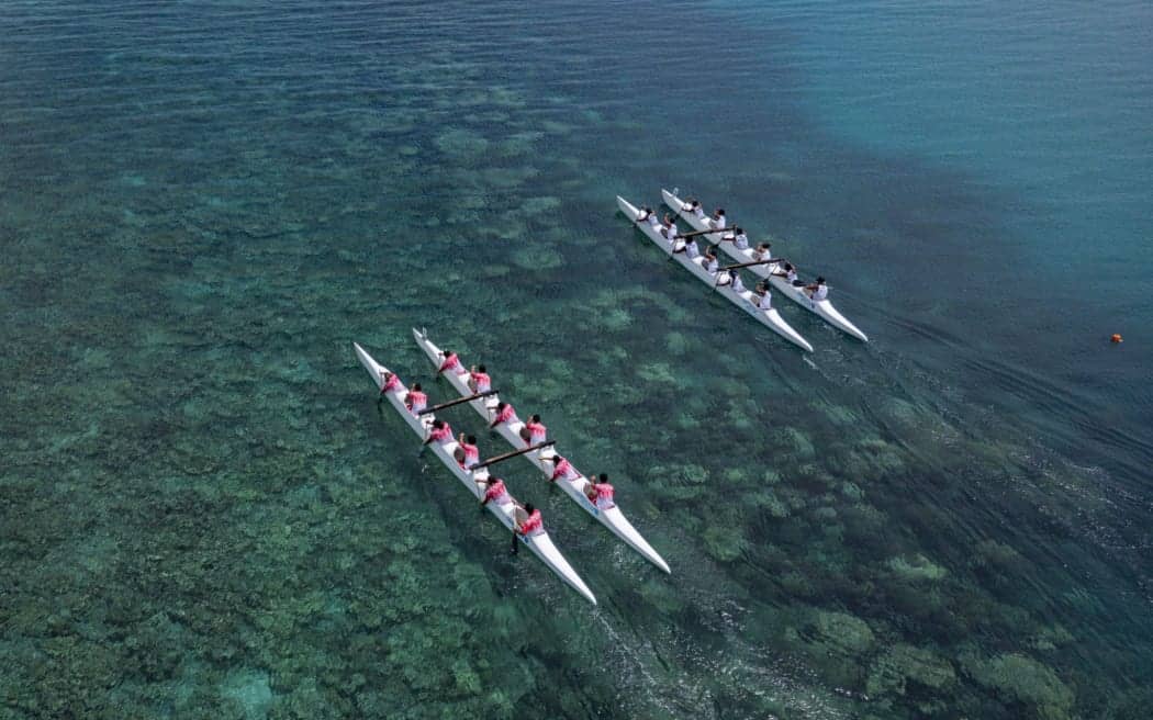 Aerial view of two outrigger canoe teams paddling side by side in clear turquoise water above a coral reef.