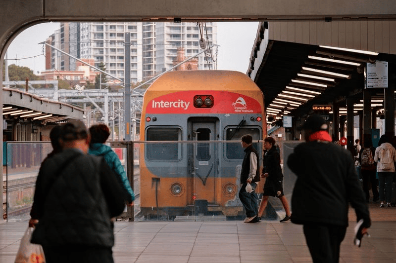 A Sydney Trains orange and silver intercity train arrives at a covered railway station platform as commuters walk along the platform. The train displays intercity branding on its front, and passengers can be seen boarding and disembarking. Modern apartment buildings are visible in the background through the station's open sides.