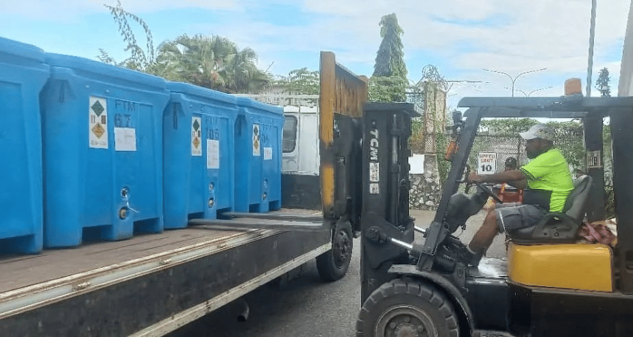 A man operating a forklift loads large blue plastic containers onto a flatbed truck. The setting appears to be an outdoor facility with trees, a stone wall, and a truck in the background.