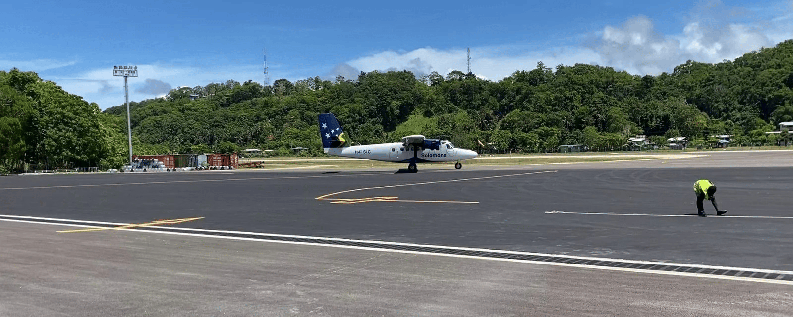A white twin-engine Solomon Airlines aircraft with blue tail featuring the national flag sits on an airport runway surrounded by dense green tropical forest, with a ground crew worker in high-visibility clothing visible on the tarmac.