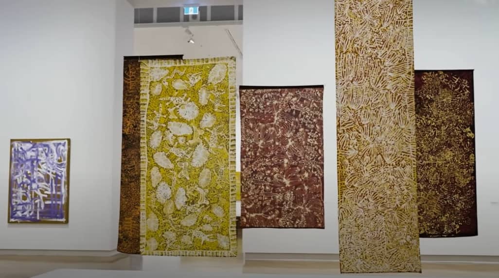 A gallery space featuring a display of large, hanging textile artworks with intricate Indigenous patterns. The textiles are predominantly in earthy tones—yellows, browns, and ochres—with organic and botanical motifs. A framed painting in purple and white hues is visible on the left wall. The textiles hang from the ceiling, creating a layered and immersive visual experience.