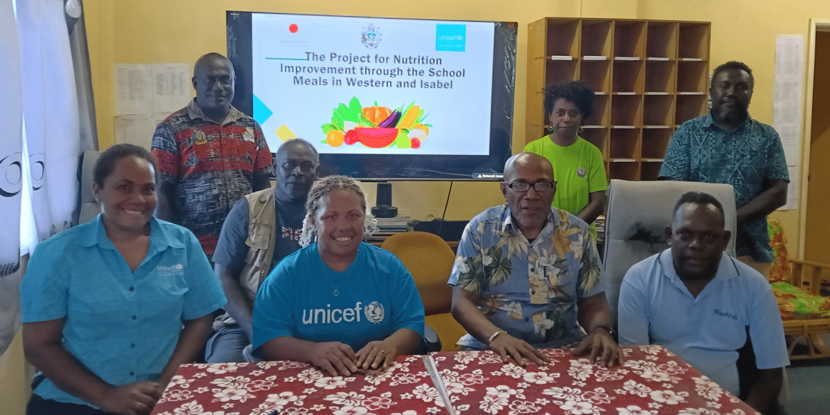 A group of eight adults is gathered in a classroom or office setting, seated and standing behind a red floral tablecloth. Most are wearing lapel pins or shirts with logos, and one individual is wearing a blue UNICEF t-shirt. In the background, a large television screen displays a colorful banner that reads, “The Project for Nutrition Improvement through the School Meals in Western and Isabel.” The room contains open cubby shelves, paperwork, and a window with curtains. The scene suggests a meeting or workshop related to a UNICEF-supported school meals initiative.