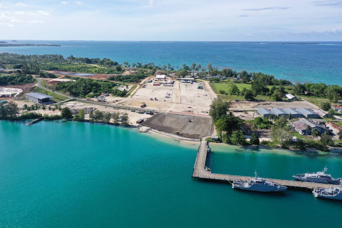 An aerial view of a coastal naval base under development, with cleared land, construction equipment, and several buildings scattered throughout the site. A long pier extends from the shore into bright turquoise water, where two naval ships are docked. The background features lush greenery and trees, with the open sea and a distant island visible under a clear sky.