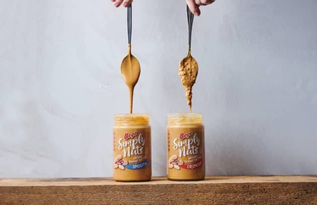 Two jars of Bega Simply Nuts peanut butter are placed on a wooden surface against a plain background. The jar on the left is labeled Smooth, and a spoon above it is dripping creamy peanut butter. The jar on the right is labeled Crunchy, with a spoon above it lifting thick, textured peanut butter with visible peanut chunks. Both spoons are held by hands from above.