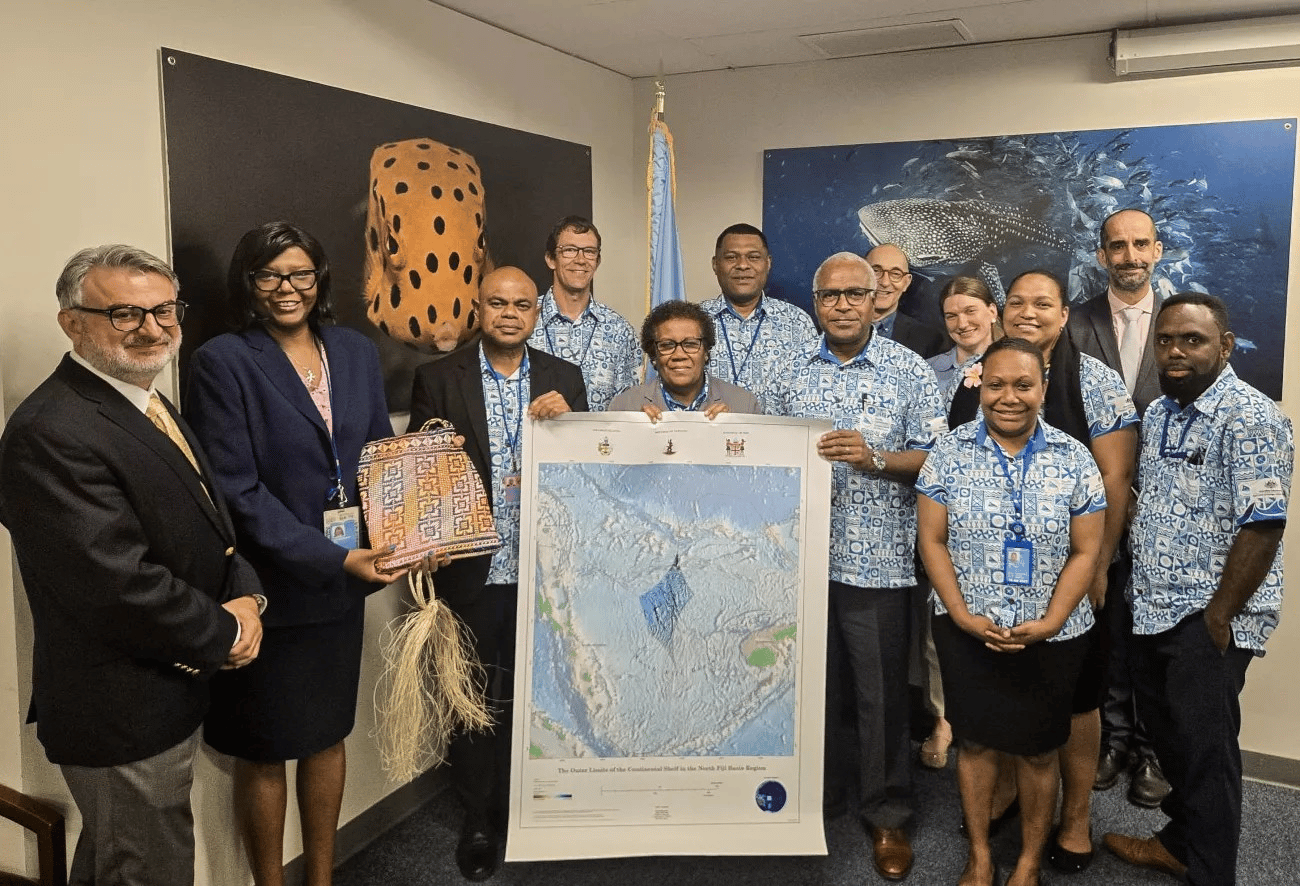 A group of people pose together in an office setting, celebrating a formal occasion. Several individuals are wearing matching blue and white Pacific-style shirts with traditional patterns. At the center, two people hold a large map titled The Outer Limits of the Continental Shelf in the North Fiji Basin Region. Another person holds a colorful woven basket adorned with a fiber handle. Behind them are two large photographs of marine life, including a spotted fish and a whale shark. The mood is professional and celebratory.
