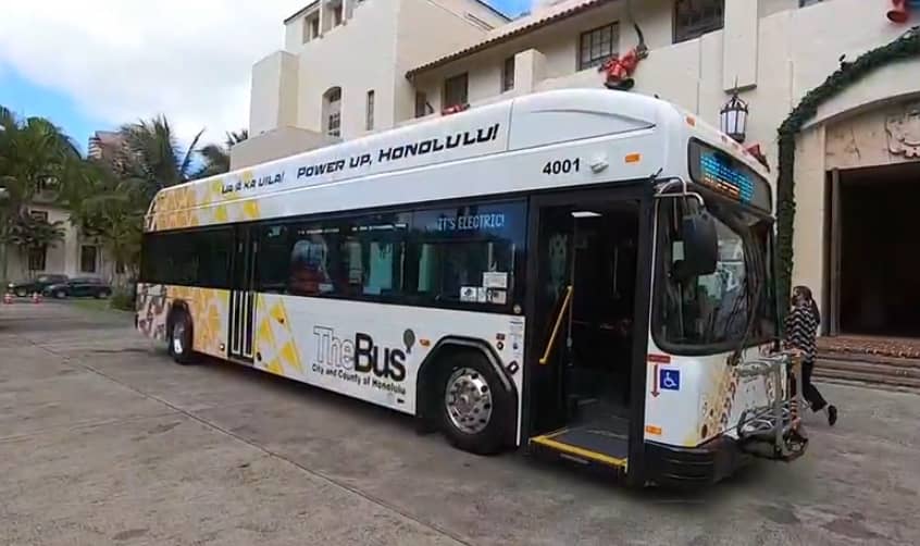 A full-length, slightly angled shot of a white city bus, operated by TheBus City and County of Honolulu, parked in front of a light-colored building with large windows and an arched entryway. The bus has POWER UP, HONOLULU! and IT'S ELECTRIC written on its side. Its rear half features a graphic design of yellow and brown patterns, resembling a floral or leaf motif. The front door of the bus is open, and a person is visible near the entrance. A ramp for accessibility is deployed from the bus.