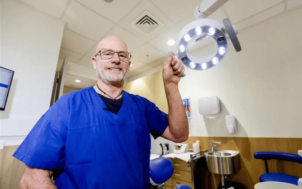 Smiling male medical professional, Dentist Dr Hugh Trengrove, wearing blue scrubs and glasses, adjusting a circular LED examination light in a clinical room with medical equipment and a sink in the background.