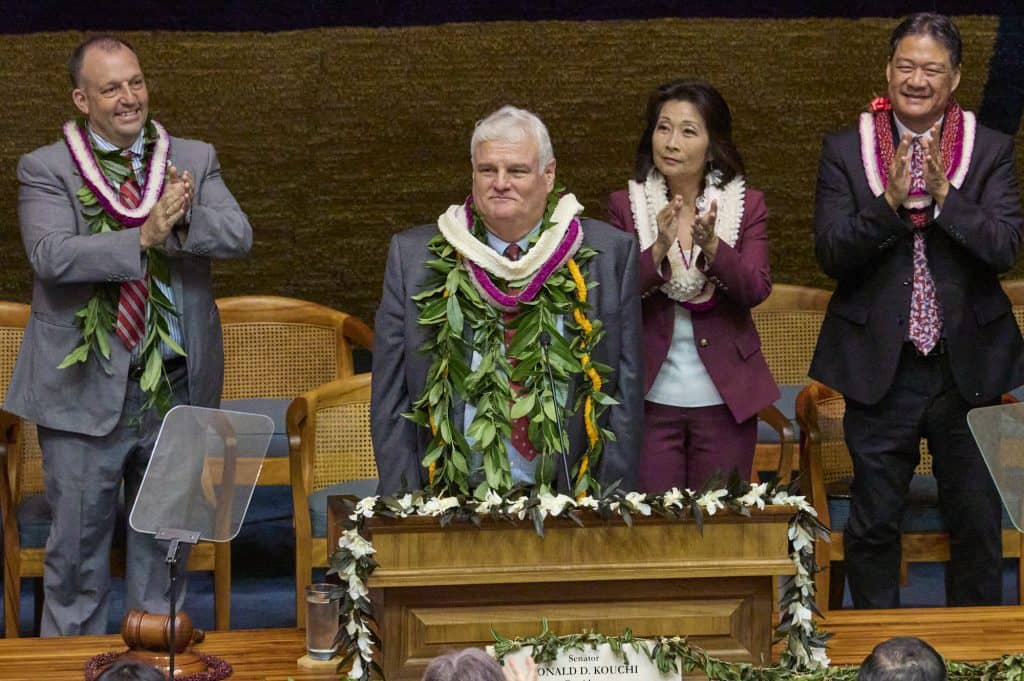 Four officials in business attire stand applauding in what appears to be a legislative chamber or formal government setting. All are wearing traditional Hawaiian lei - the men wear maile leaf lei while the woman wears white plumeria lei. The central figure, an older man with white hair in a dark suit, wears an elaborate maile lei and stands behind a wooden podium decorated with white plumeria flowers. A nameplate reading RONALD D. KOUCHI is visible on the podium. The setting features wooden furniture and blue carpeting typical of a state capitol building.